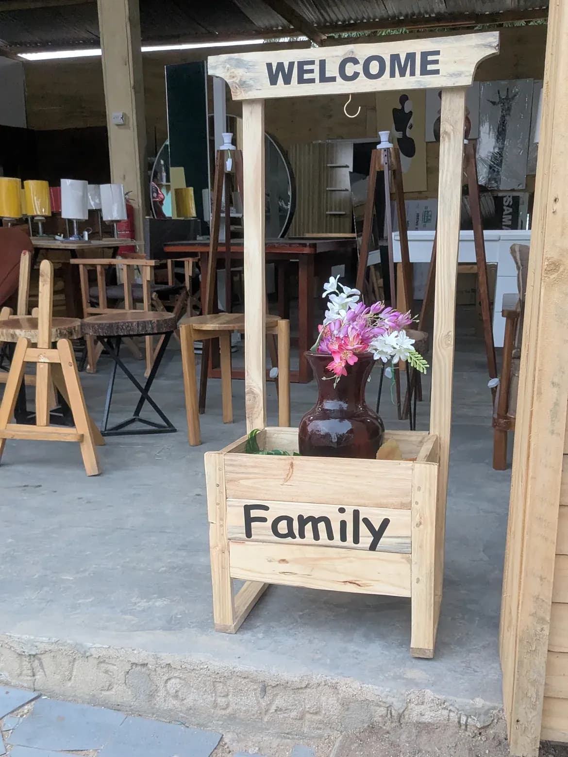 A wooden 'Welcome' sign above a 'Family' planter with pink flowers at The Artist showroom entrance.